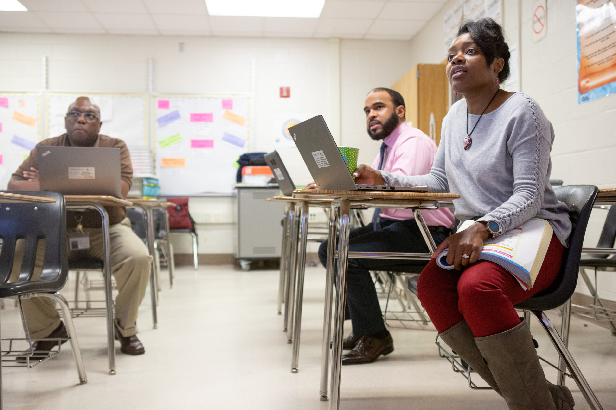 An image of three educators of color who are sitting inside a classroom at desks. They each have a laptop in front of them.