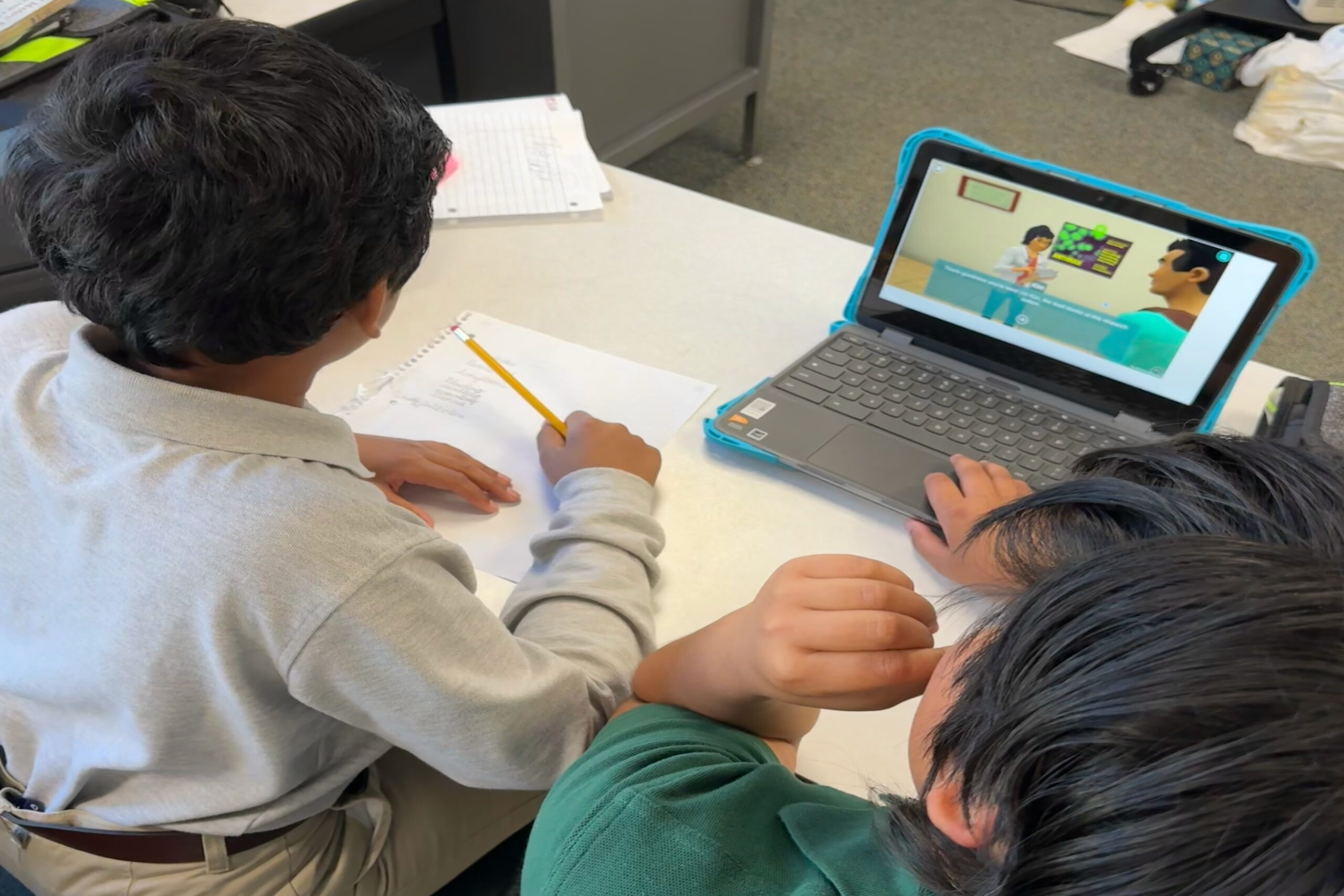 An image of two male students taking notes on paper, while they look at a laptop on the desk in front of them