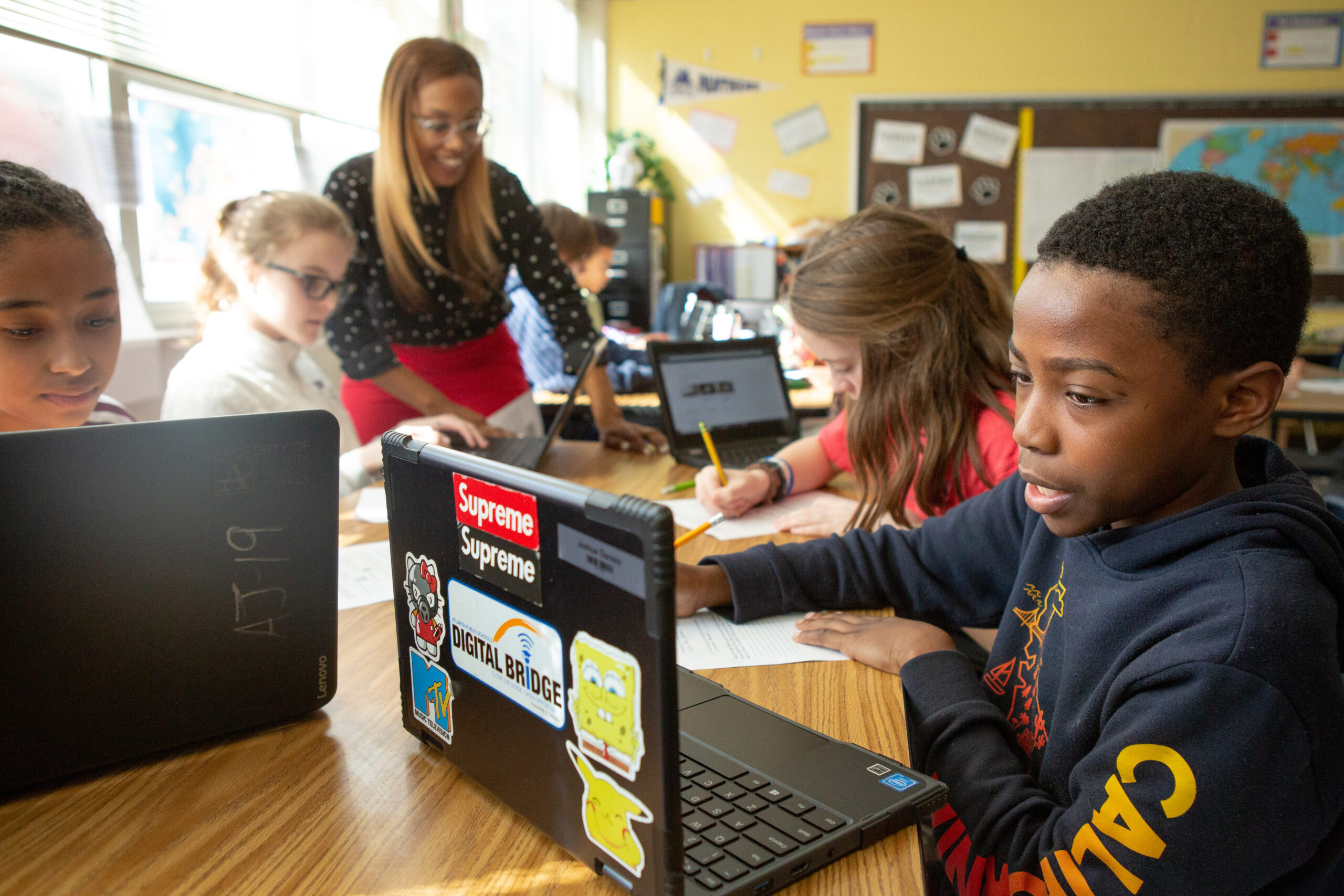An image of a diverse group of students working collaboratively at a classroom table using laptops. A smiling teacher stands nearby, offering guidance. One boy in the foreground is focused on his laptop, which is decorated with colorful stickers including “Supreme,” “Digital Bridge,” and cartoon characters. Other students are writing or typing, and educational posters and a world map are visible on the classroom walls.