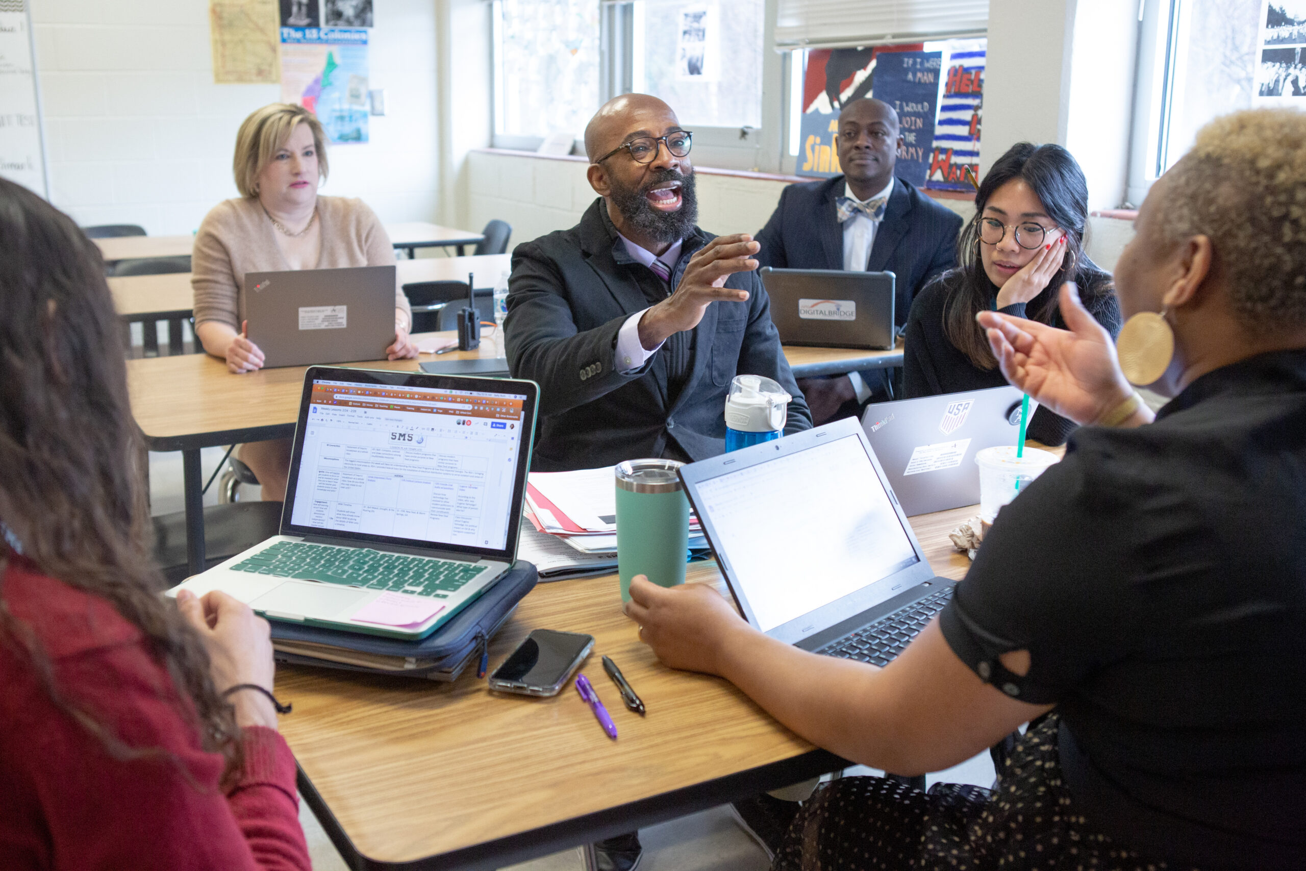 An image of six educators, of varied race and genders, sitting in a classroom discussing lesson plans