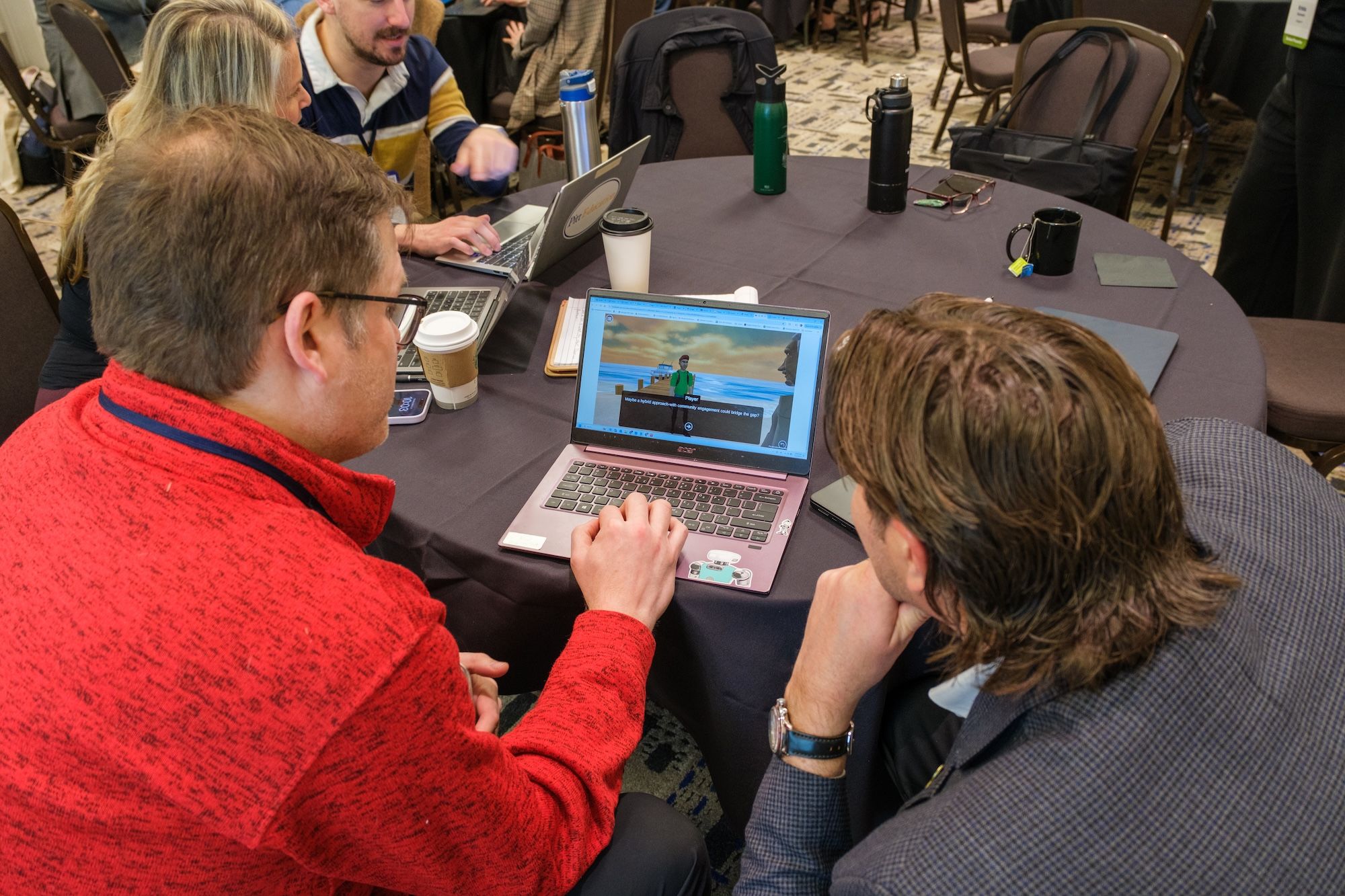 Two men sit at a table looking at SceneCraft on a laptop.