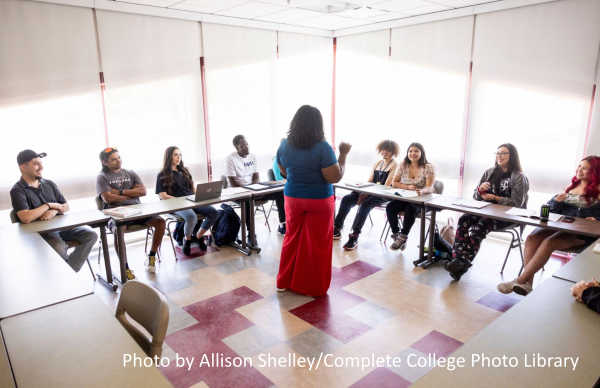 Black female instructor speaking to a multiracial group of students during a class in a sunny classroom on a college campus.