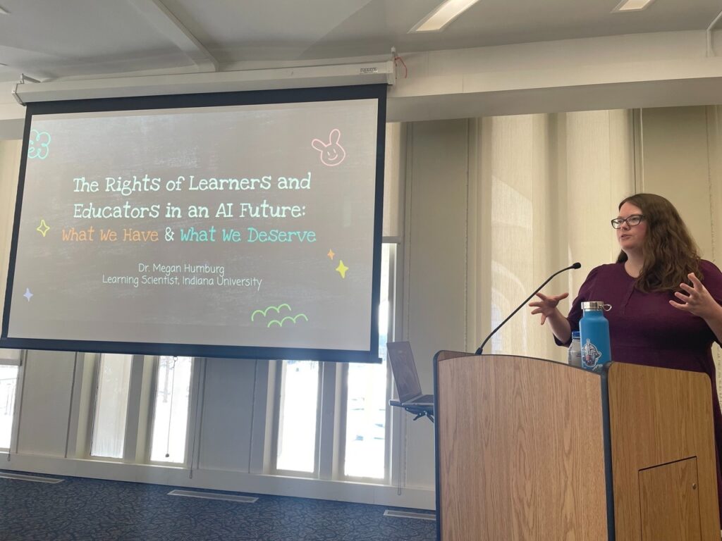 A woman standing at a podium talking, with a powerpoint behind her that reads "The Rights of Learners and Educators in an AI Future: What we have and what we deserve"